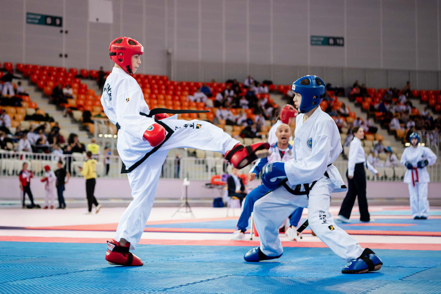 The scene is set in a large indoor sports arena, where several people are engaged in martial arts training. There are at least twelve individuals present, with some of them wearing uniforms and practicing their moves on the blue mats.nnA man can be seen kicking another person's leg during the training session, while other participants watch and learn from each other. The atmosphere is lively as everyone focuses on improving their skills in various martial arts techniques.